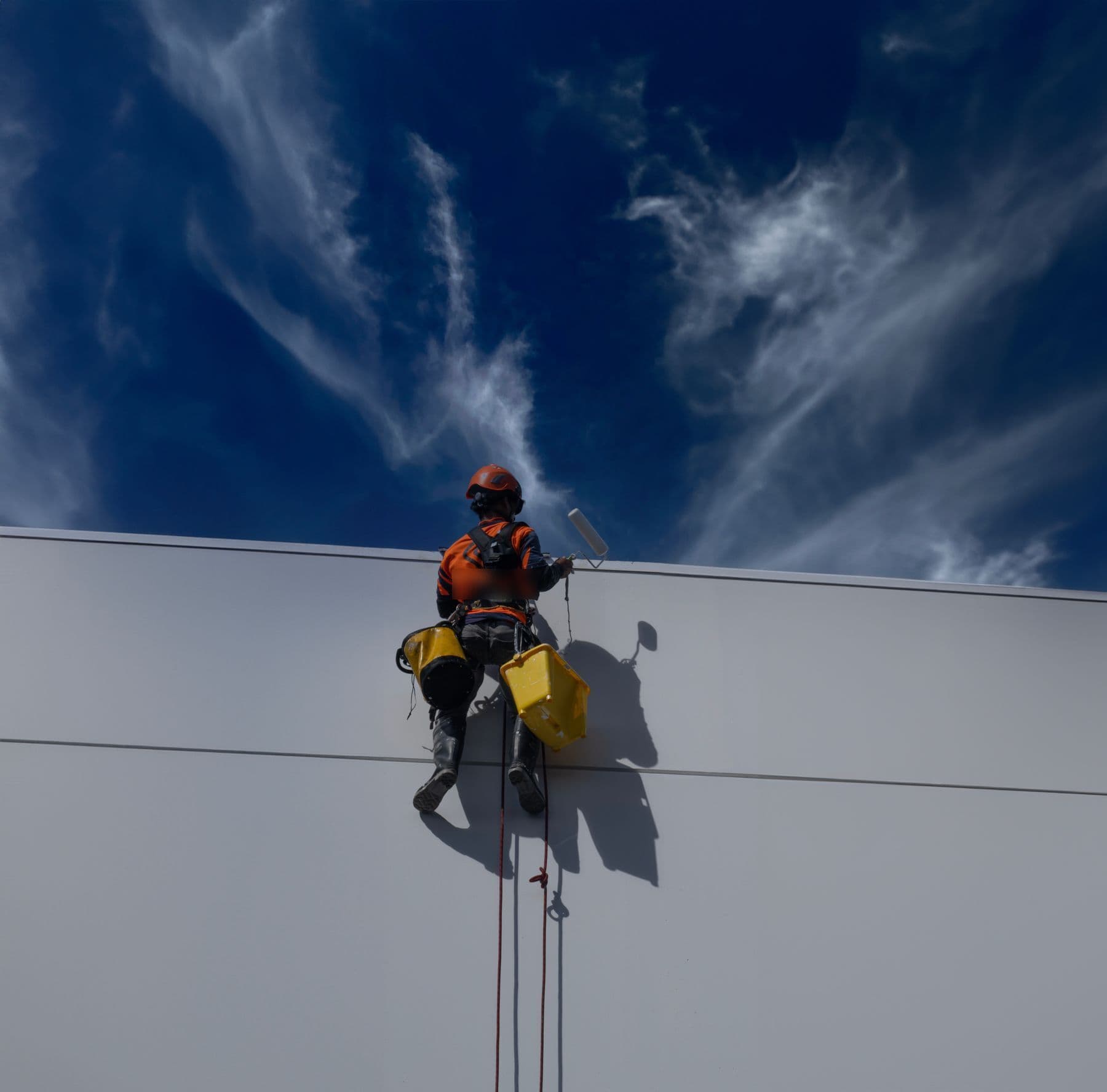A worker hanging on ropes beside a white wall, rolling paint near the roofline under a deep blue sky.