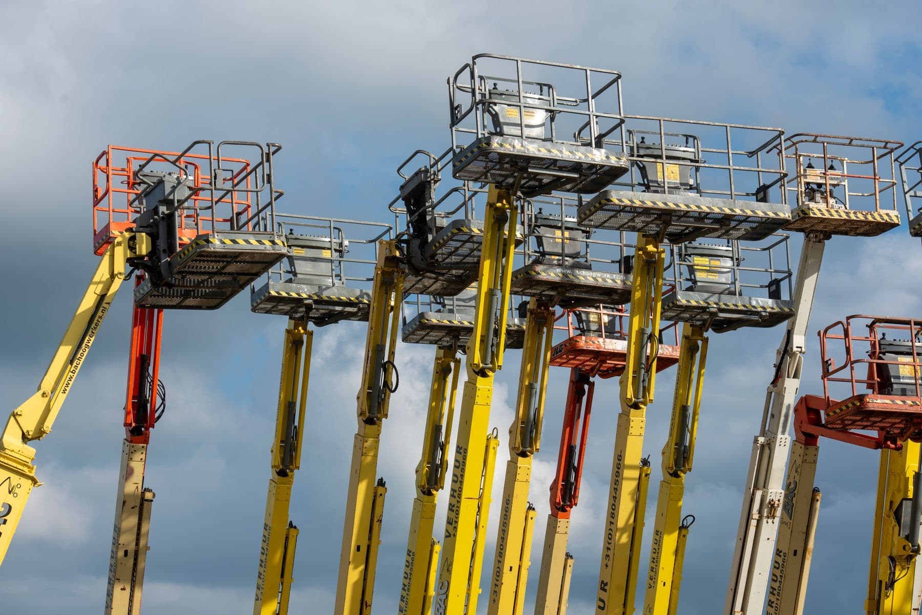 Several yellow and orange boom lifts raised against a cloudy blue sky.