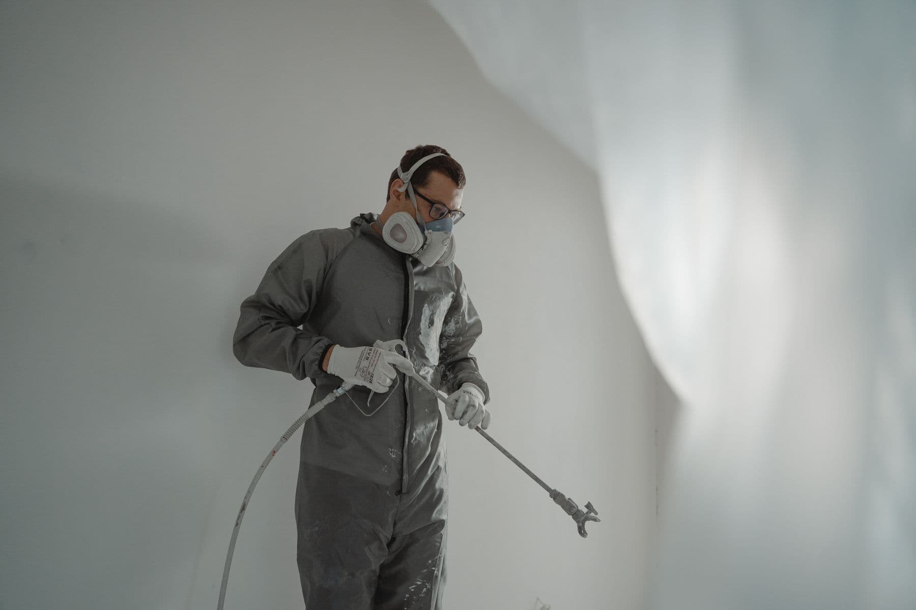 A worker in a gray protective suit and respirator holding an airless spray wand in a white room.