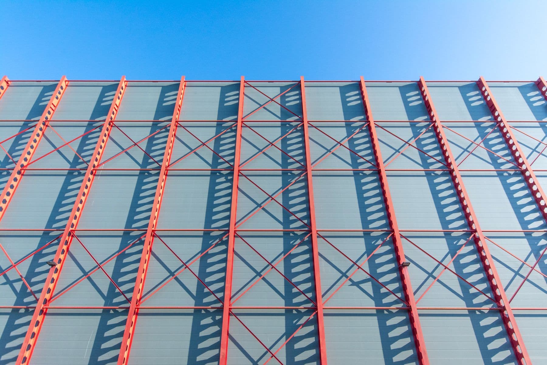 Red steel beams crossing a glass and metal building facade against a blue sky.